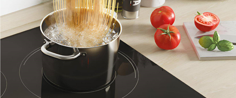 Na płycie indukcyjnej znajduje się garnek z gotującą się wodą i makaronem. Obok na blacie leżą pomidory i liście bazylii.