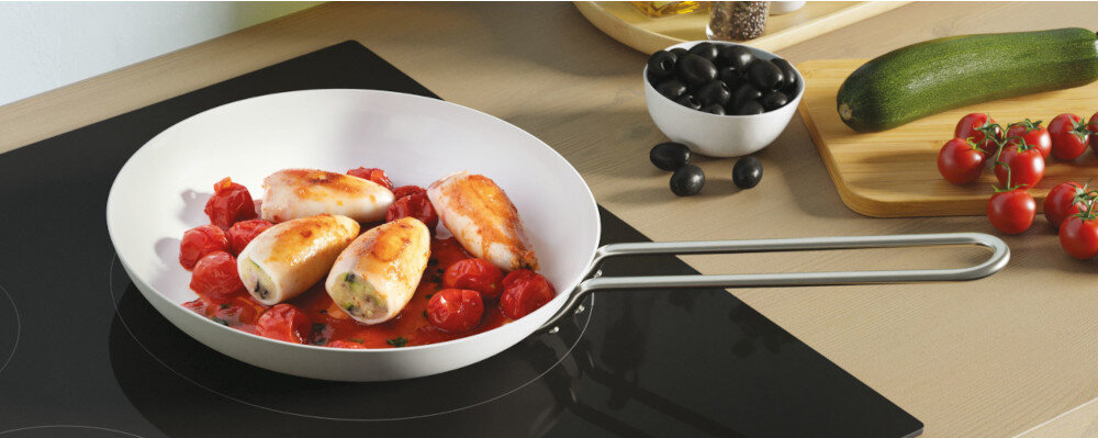 Na płycie indukcyjnej znajduje się patelnia z potrawą złożoną z pomidorów i nadziewanych roladek. Obok leży deska z cukinią i pomidorkami koktajlowymi oraz miska z czarnymi oliwkami.