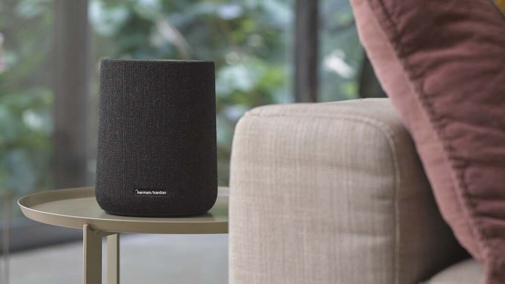Czarny głośnik z logo \'Harman Kardon\' stoi na okrągłym stoliku obok beżowej sofy. W tle widoczne są rozmyte zielone rośliny za oknem.