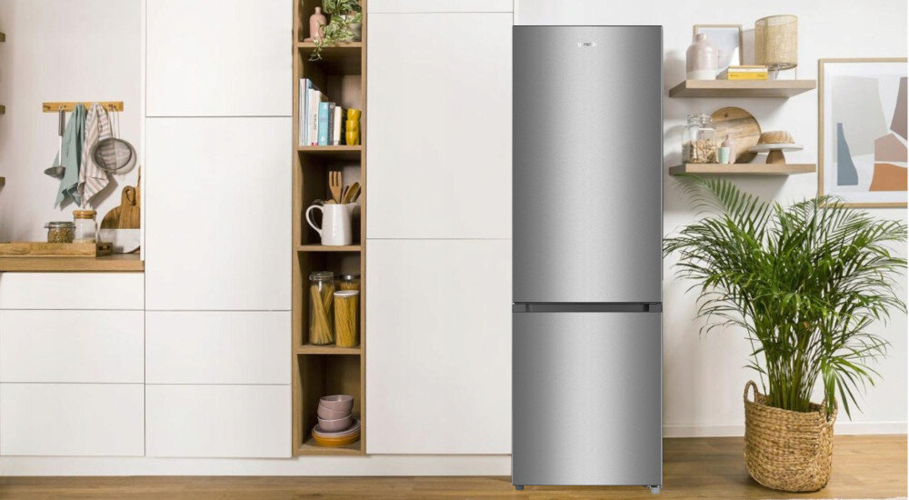 Srebrna lodówka stoi w nowoczesnej kuchni z białymi szafkami i drewnianymi półkami. Obok lodówki znajduje się duża roślina w plecionym koszu.