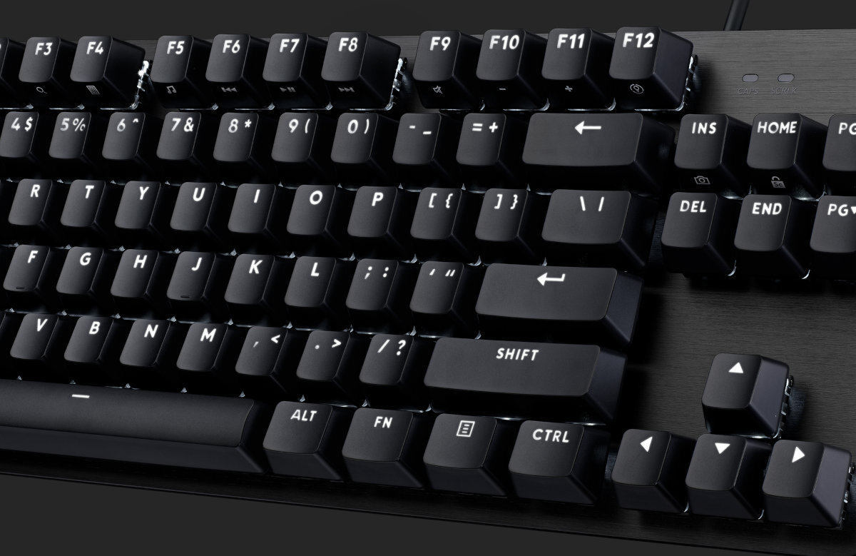 Czarna klawiatura z białymi oznaczeniami na klawiszach, w tym klawisze funkcyjne F1 do F12 oraz klawisze strzałek. Widoczne są także klawisze z symbolami specjalnymi, takie jak @, #, $, %, ^, &, *, (, ), _, +, {, }, |, :, \