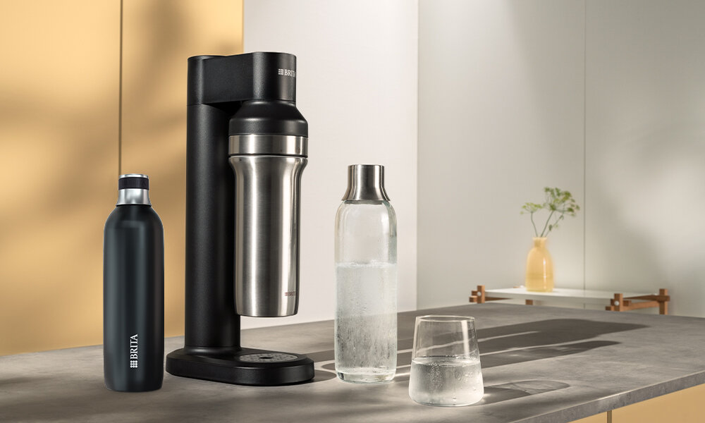 Na blacie kuchennym stoi czarny saturator BRITA SodaTRIO oraz butelka z wodą gazowaną i szklanka. Obok znajduje się czarna butelka z logo BRITA.
