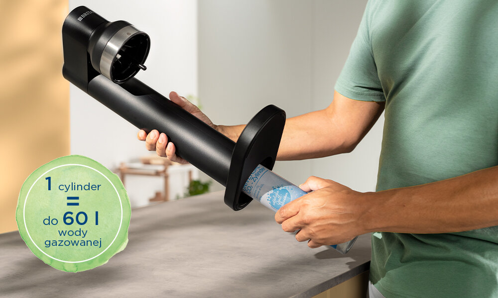 Osoba trzyma czarny saturator z cylindrem w rękach. Na obrazie widoczny jest tekst: \'1 cylinder = do 60 l wody gazowanej\'.