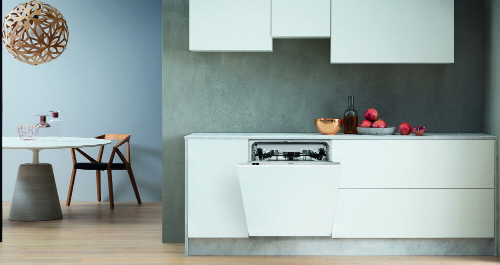 Nowoczesna kuchnia z otwartą zmywarką w zabudowie, na blacie stoją owoce i butelka. W tle widoczny jest stół z krzesłem oraz dekoracyjna lampa sufitowa.