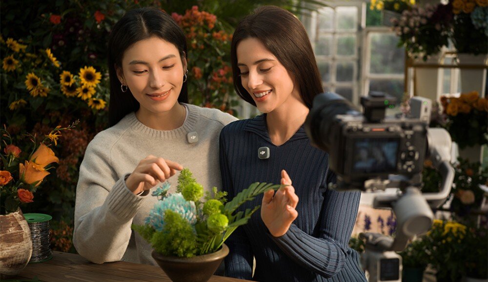Nadajnik DJI Mic Mini 2 dwie kobiety w szklarni przy stole z kwiatami i aparatem na statywie, obie mają przypięte małe, szare mikrofony, akcesoria