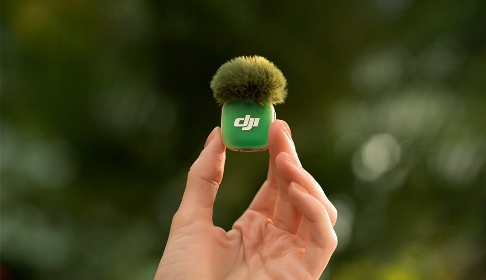 Nadajnik DJI Mic Mini 2 dłoń trzymająca mały, zielony mikrofon z nałożoną futrzaną osłoną przeciwwietrzną na rozmytym tle zieleni, cechy, przeznaczenie, zalety