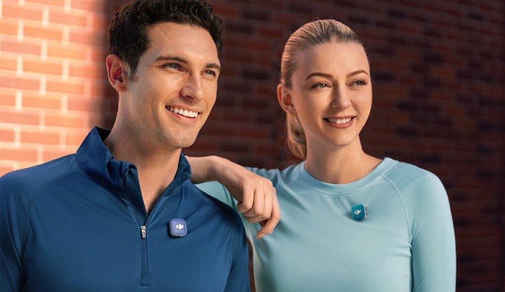 Nadajnik DJI Mic Mini 2 uśmiechnięta para w sportowych strojach (mężczyzna w niebieskiej bluzie, kobieta w jasnoniebieskiej) z przypiętymi mikrofonami, stojąca przed ceglaną ścianą, tryby działania, redukcja szumów, nagrywanie