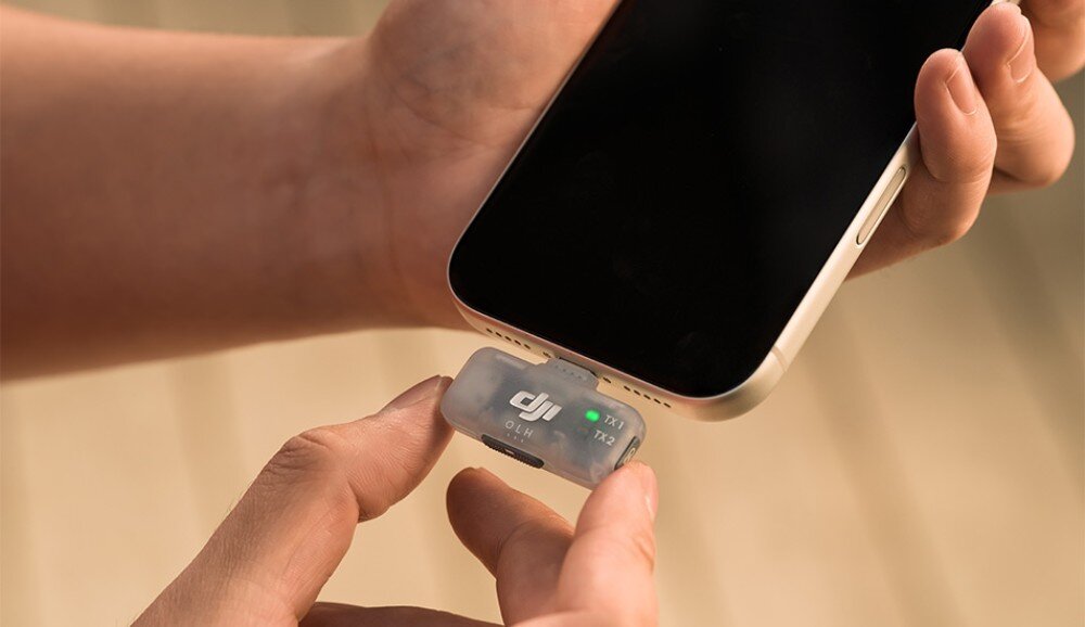 Nadajnik DJI Mic Mini 2 zbliżenie na dłonie podłączające mały, przezroczysty odbiornik z napisem 'TX1 TX2' do portu USB-C smartfona, kompatybilność, podłączenie, łączność