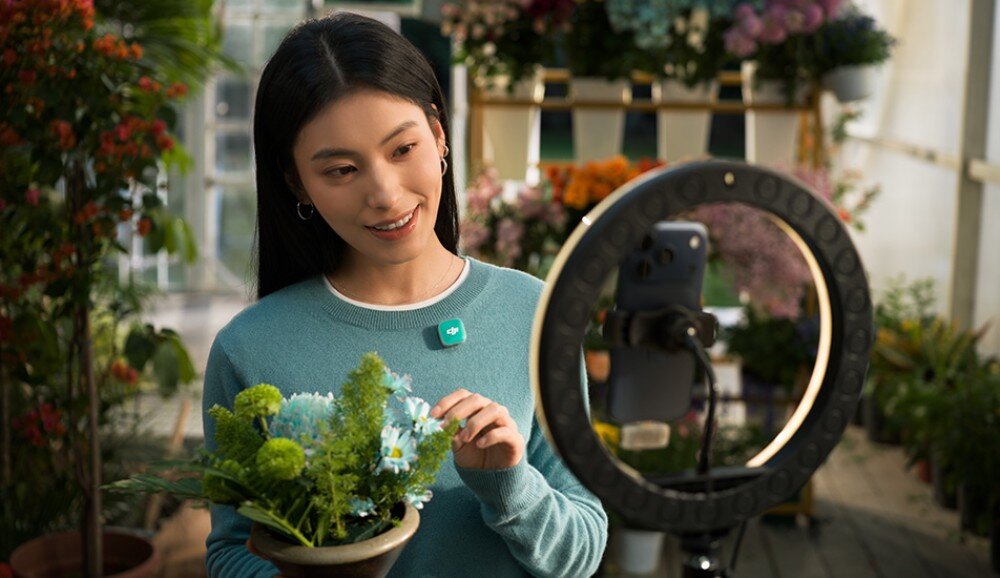 System bezprzewodowy DJI Mic Mini 2 Kobieta w błękitnym swetrze z przypiętym morskim mikrofonem uśmiecha się podczas nagrywania wideo telefonem umieszczonym w lampie pierścieniowej. W dłoniach trzyma doniczkę z kwiatami, a wokół niej widać wnętrze kwiaciarni, akcesoria