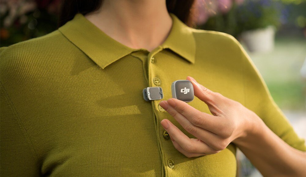 System bezprzewodowy DJI Mic Mini 2 Zbliżenie na dłoń kobiety w zielonej koszulce polo, która przykłada mały, szary, kwadratowy nadajnik do magnetycznego klipsu przymocowanego przy guzikach ubrania, nadajnik, waga, mocowanie, etui
