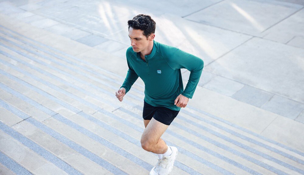 System bezprzewodowy DJI Mic Mini 2 Mężczyzna w zielonej bluzie sportowej z przypiętym mikrofonem biegnie po szerokich, kamiennych schodach na zewnątrz budynku. Zdjęcie wykonane z perspektywy górnej, ukazujące dynamikę ruchu, specyfikacja, jakość audio, funkcje