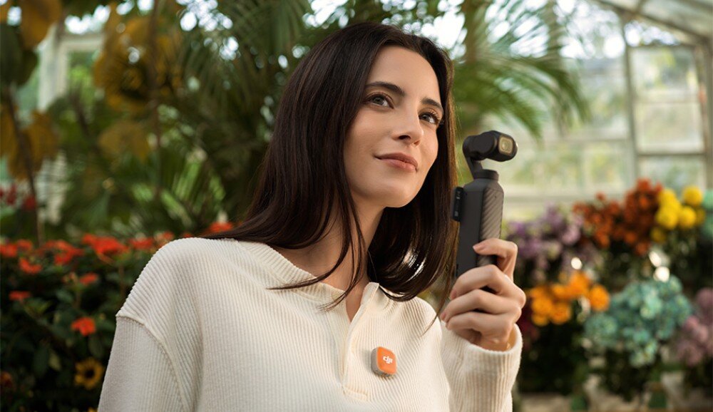 System bezprzewodowy DJI Mic Mini 2 Kobieta w białym swetrze trzyma przed sobą małą kamerę na gimbalu, mając jednocześnie przypięty do ubrania pomarańczowy nadajnik mikrofonowy. Postać znajduje się w szklarni pełnej roślin i kwiatów, aplikacja, funkcje, kompatybilność