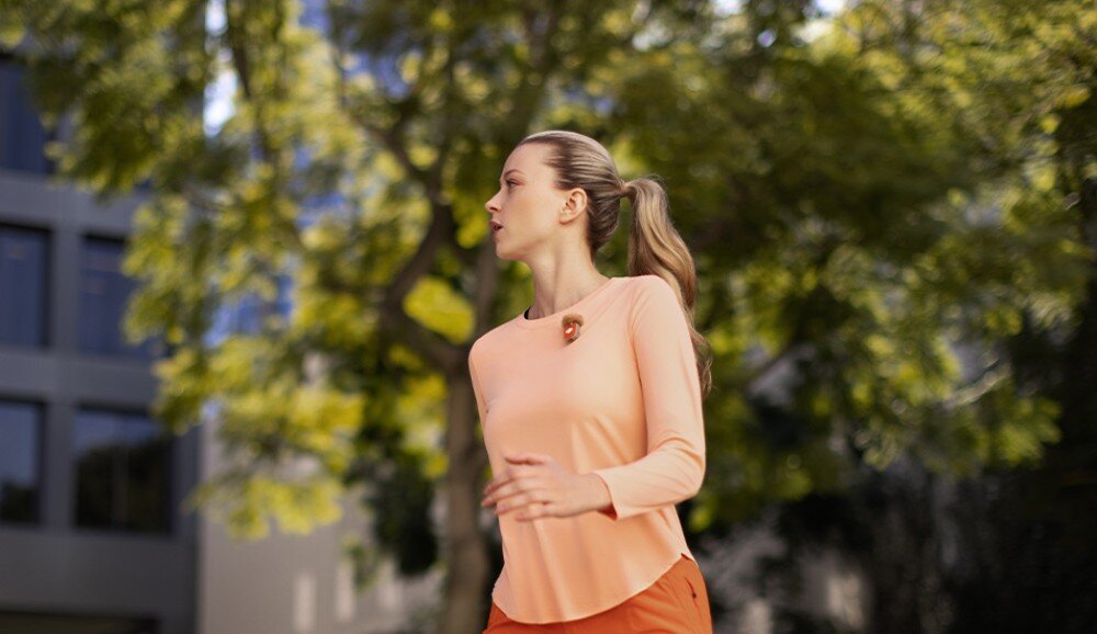 System bezprzewodowy DJI Mic Mini 2 Kobieta w pomarańczowym stroju sportowym biegnie przez park na tle rozmytych drzew i budynków. Do jej koszulki przypięty jest mikrofon z założoną brązową osłoną przeciwwiatrową typu 'deadcat', zasięg, sygnał, czas pracy, ładowanie