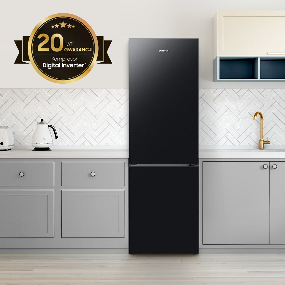 Czarna lodówka SAMSUNG stoi w nowoczesnej kuchni z szarymi szafkami i białymi płytkami na ścianie. Na ścianie widoczny jest emblemat z napisem \'20 lat gwarancji Kompresor Digital Inverter\'.