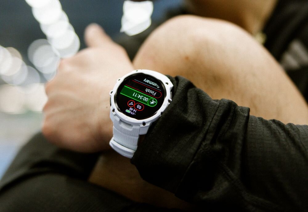 Osoba nosi biały zegarek sportowy z wyświetlaczem pokazującym czas 00:34:11 i inne dane treningowe. Zegarek ma okrągły kształt i jest umieszczony na nadgarstku ubranym w ciemną odzież.