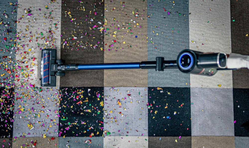 Odkurzacz z niebieską rurą czyszczący dywan w kratkę, pokryty kolorowymi konfetti.