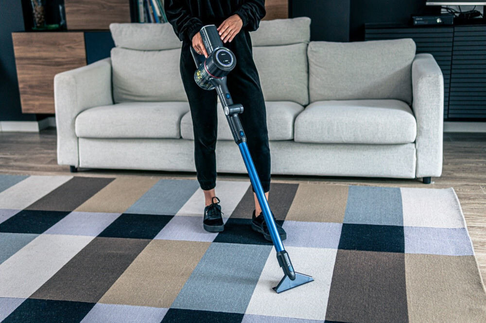Osoba używa odkurzacza pionowego na kolorowym dywanie w salonie z jasną sofą w tle. Odkurzacz ma niebieską rurę i przezroczysty pojemnik na kurz.