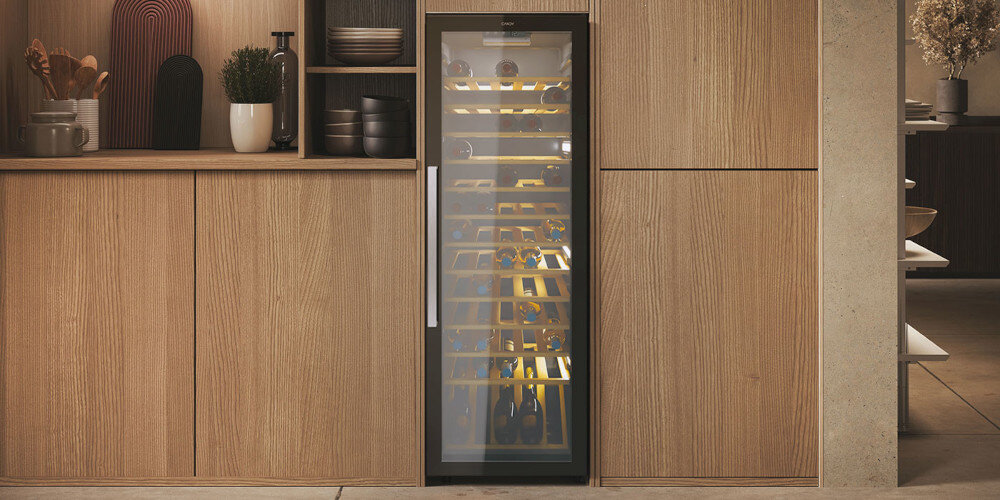 Chłodziarka do wina z przeszklonymi drzwiami, wbudowana w drewnianą zabudowę kuchenną. Wewnątrz widoczne są półki z butelkami wina.