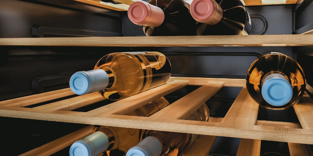 Wnętrze chłodziarki do wina z drewnianymi półkami, na których leży kilka butelek wina z różnokolorowymi zakrętkami. Butelki są ułożone poziomo, a wnętrze jest oświetlone ciepłym światłem.