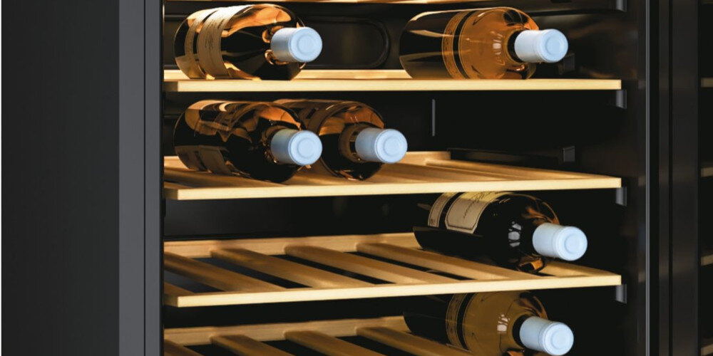 Wnętrze chłodziarki do wina z drewnianymi półkami, na których leży kilka butelek wina z białymi korkami. Chłodziarka ma ciemne wnętrze i jest oświetlona ciepłym światłem.