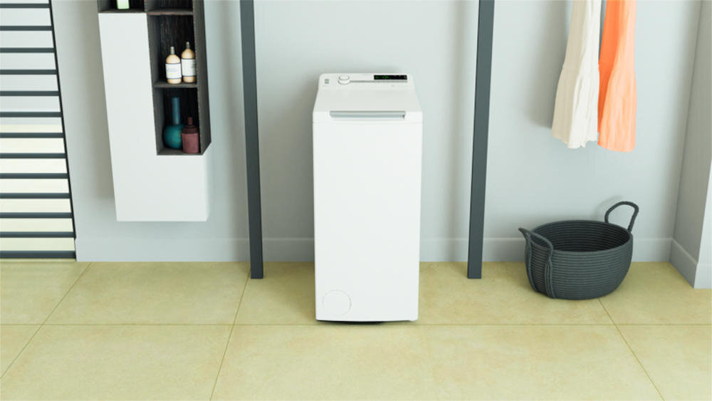 Na obrazie znajduje się biała pralka ładowana od góry, ustawiona w jasnym pomieszczeniu. Obok pralki widoczny jest kosz oraz szafka z kosmetykami.