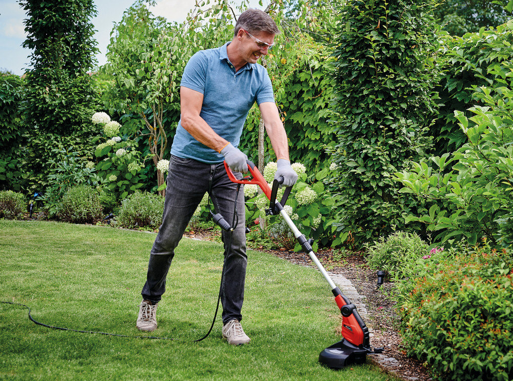 Osoba używa podkaszarki elektrycznej na trawniku w ogrodzie. W tle widoczne są krzewy i drzewa.