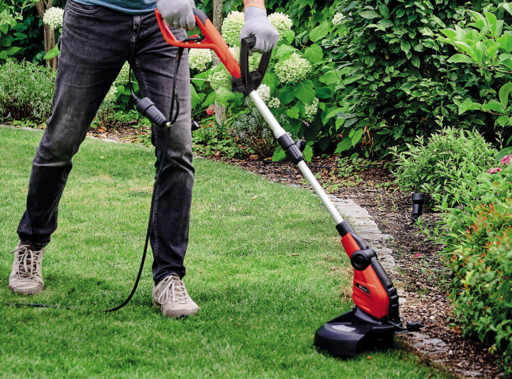 Osoba używa czerwono-czarnej podkaszarki elektrycznej na trawniku w ogrodzie. W tle widoczne są zielone krzewy i kwiaty.