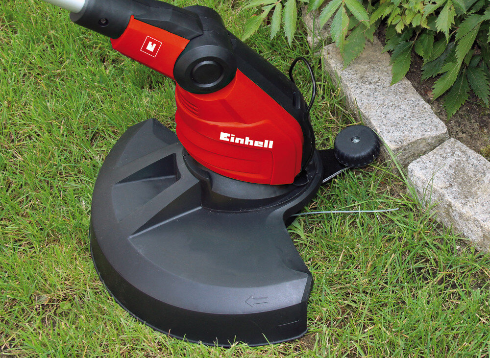 Na obrazie widoczna jest czerwona podkaszarka elektryczna z czarną osłoną, leżąca na trawie obok kamiennej krawędzi. Na urządzeniu znajduje się logo \'Einhell\'.