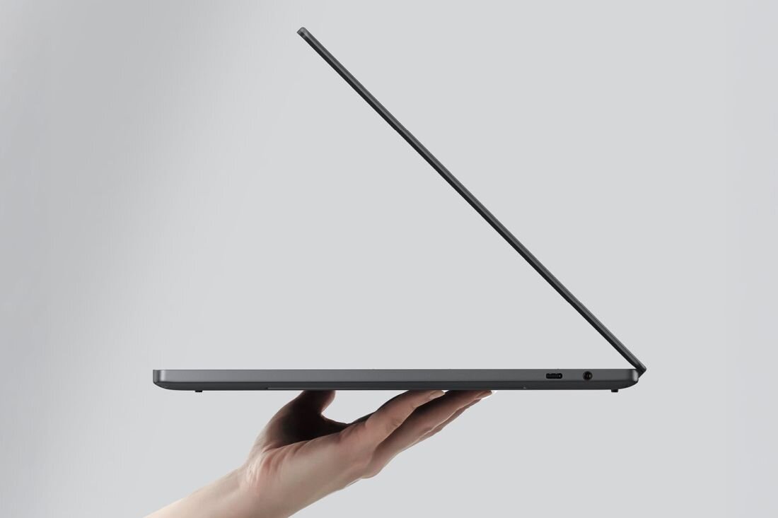 Laptop w kolorze ciemnoszarym jest trzymany w jednej ręce, z ekranem częściowo otwartym. Na obudowie widoczne są porty USB i gniazdo słuchawkowe.
