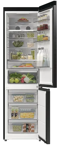 Otwarta lodówka z różnymi produktami spożywczymi na półkach, w tym warzywa, owoce, napoje i pojemniki z jedzeniem. W dolnej części znajdują się szuflady z zamrożonymi produktami.