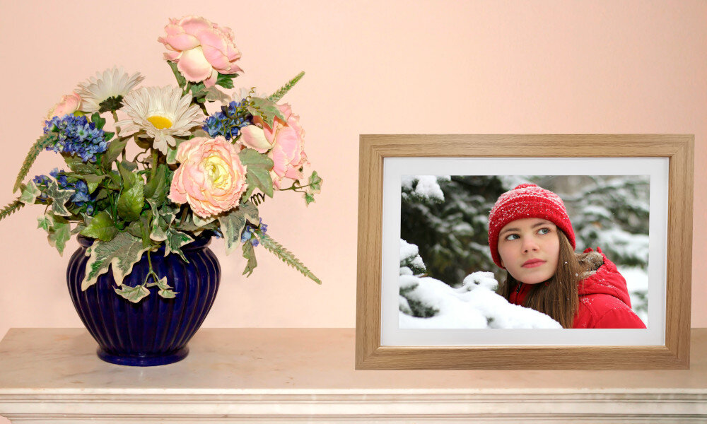 Na półce stoi niebieski wazon z kolorowymi kwiatami obok cyfrowej ramki z drewnianą obwódką, w której wyświetlane jest zdjęcie osoby w czerwonej czapce i kurtce na tle zaśnieżonego krajobrazu.