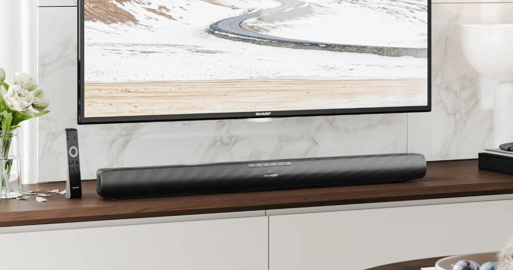 Na drewnianej półce znajduje się czarny soundbar z logo SHARP, przed telewizorem wyświetlającym zimowy krajobraz. Obok soundbara leży pilot, a po lewej stronie stoi wazon z białymi kwiatami.